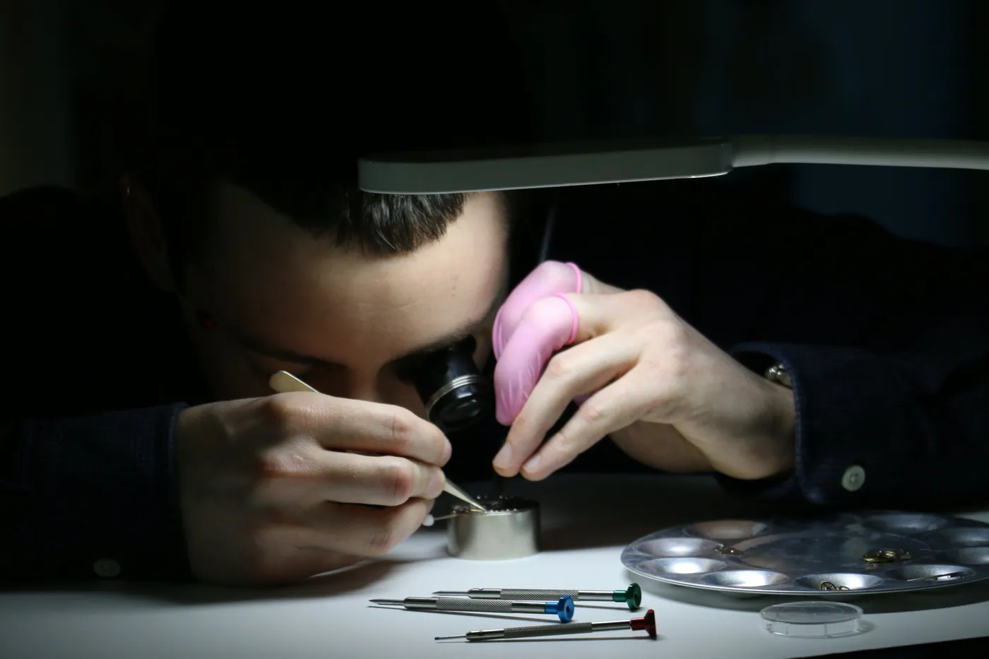A participant with a loupe and finger cots works on a movement with tweezers under a bench lamp