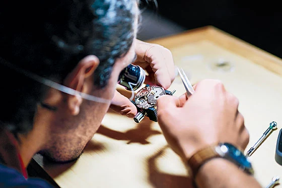 A watchmaker with a loupe works on a movement held in a movement holder at a bench in the HSNY workshop